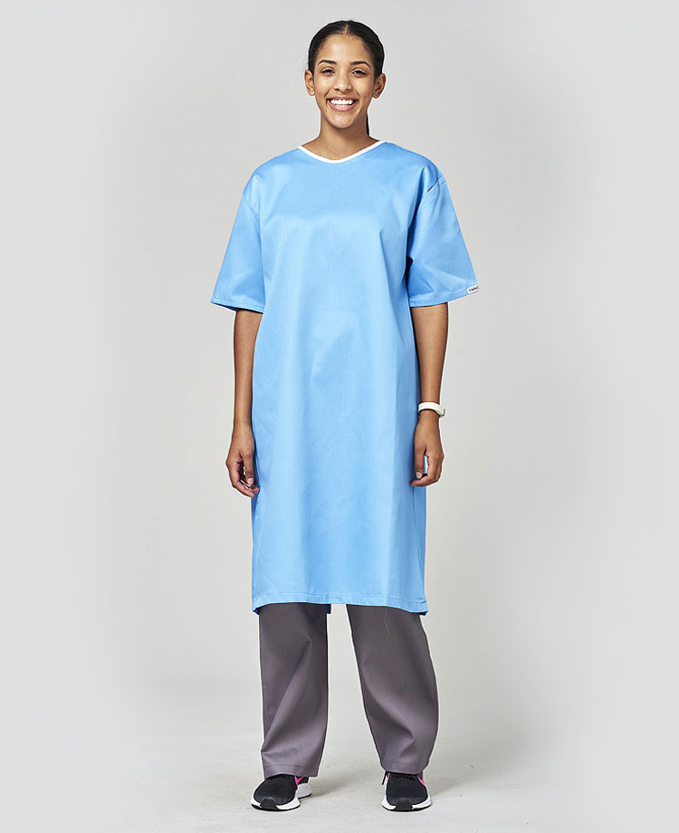 A woman wearing a light blue hospital gown with short sleeves, smiling and standing confidently. The gown features a simple, comfortable design, ideal for medical settings.