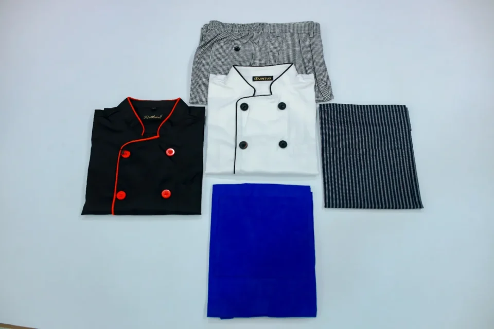 Chef uniforms set including white coat, black coat with red piping, chef trousers, and apron by Reethatex Trivandrum
