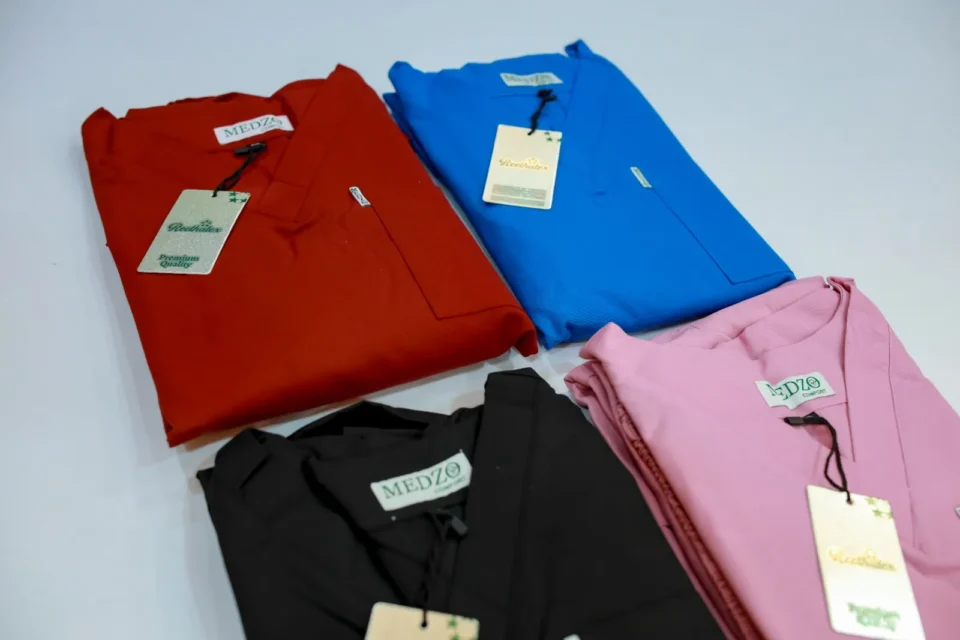Folded medical scrub tops in red, royal blue, black, and pink with brand tags by Reethatex Trivandrum